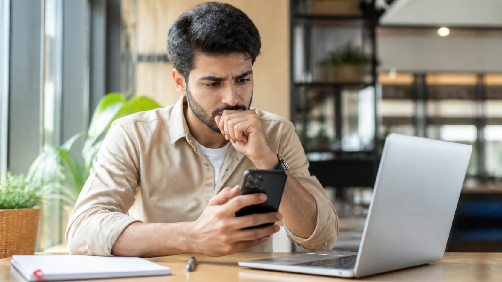 Young professional man cautiously checking a job message on his phone at a laptop desk in a bright office
